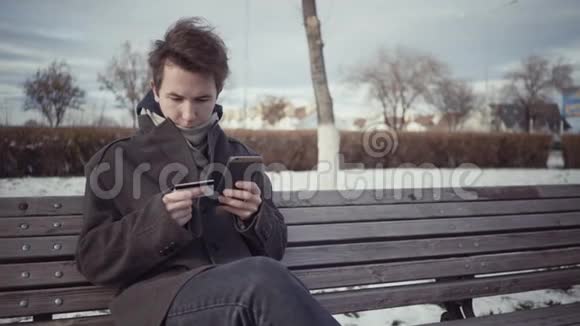 冬季坐在长凳上用信用卡和智能手机在线支付的男士视频的预览图