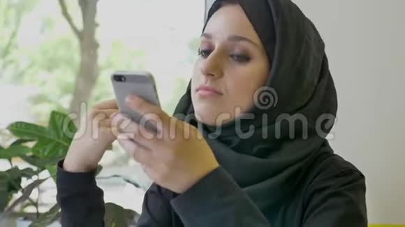 年轻漂亮的穆斯林妇女戴着头巾坐在咖啡馆里拿着电话打着电话想着决心视频的预览图