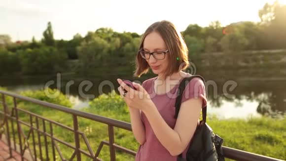 学生女孩用智能手机站在桥上的户外希普斯特在电话上浏览互联网发短信和视频的预览图