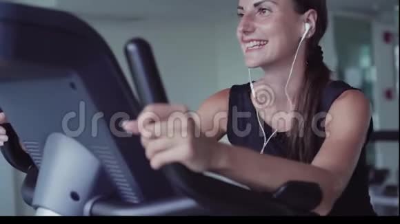 在减肥女性健身中心锻炼自行车有氧运动女性在耳机上听音乐运动员视频的预览图