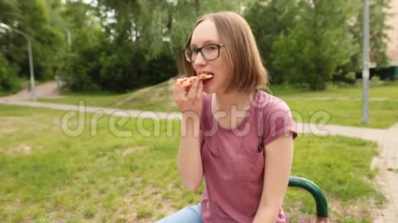 戴眼镜的滑稽时髦女孩坐在长凳上吃一片比萨饼视频的预览图