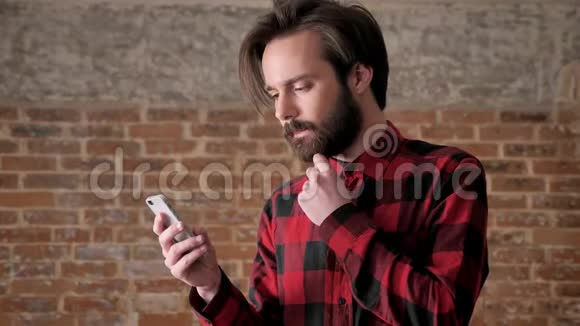 年轻的英俊男子带着胡须手表在智能手机上放大照片交流概念砖背景视频的预览图