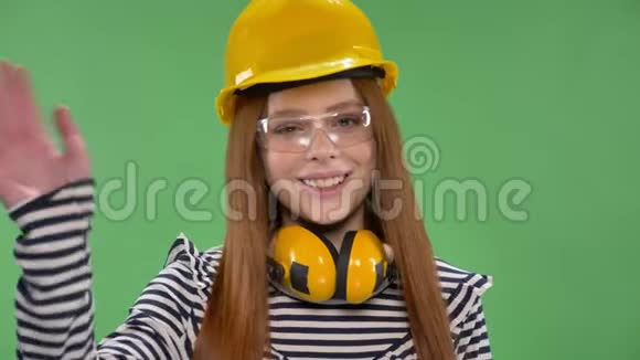 年轻的红发少女戴着建筑头盔耳机护目镜挥动她的手打招呼视频的预览图