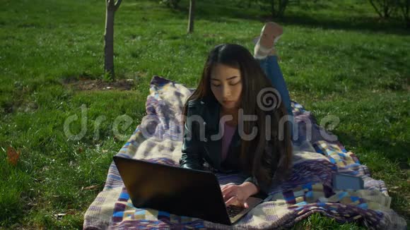 公园里躺在草坪上用笔记本电脑打字的时髦女孩视频的预览图