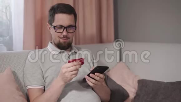 成年男子坐在家里的沙发上将卡片数据放入智能手机应用程序视频的预览图