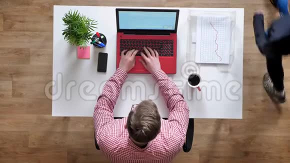 戴眼镜的年轻人在笔记本电脑上工作和打字有人四处走动顶头坐在桌子后面喝咖啡视频的预览图