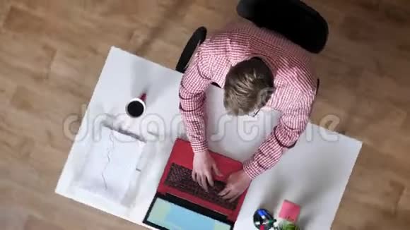 戴眼镜的年轻人在手提电脑上工作和打字顶镜头坐在桌子后面喝咖啡相机在周围盘旋视频的预览图