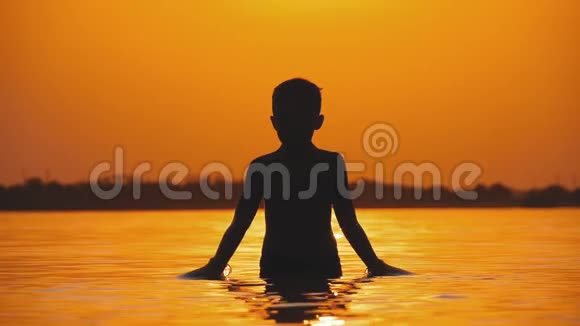 日落时分男孩的剪影举起双手创造水的飞溅慢动作视频的预览图