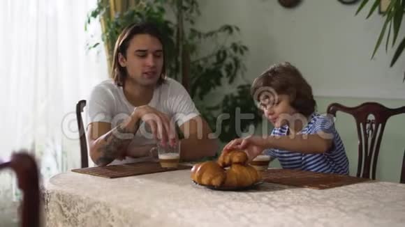 两个兄弟坐在厨房的桌子上吃着牛角面包和卡布奇诺兄弟关系视频的预览图