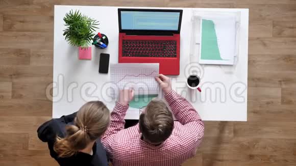 戴眼镜的年轻男子和拿着文件说话和指指点点的女人上面有照片桌子上有咖啡和笔记本电脑视频的预览图