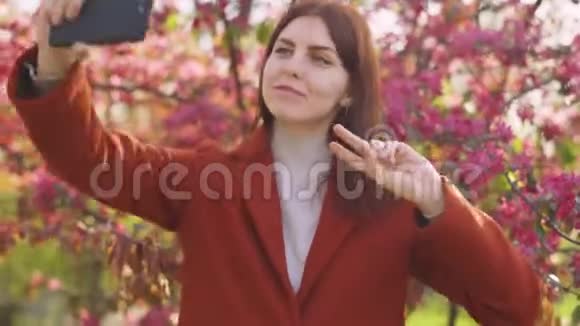 年轻迷人的红发女人在智能手机上拍摄樱花或樱花春花的背景自拍视频的预览图