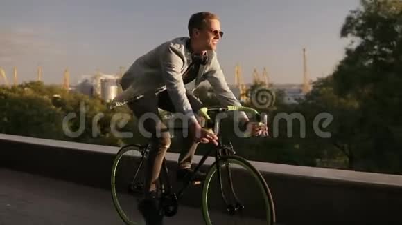 快乐的年轻微笑男子戴着太阳镜骑自行车在早晨公园或林荫大道年轻人的慢动作视频的预览图