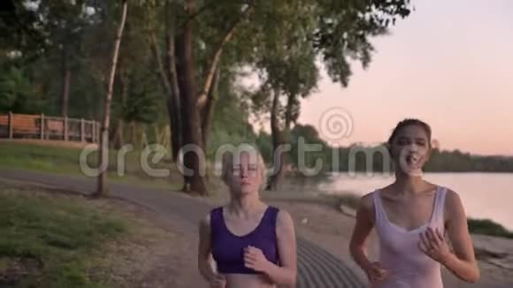日落时分两位梳着马尾辫的年轻漂亮的女跑步者在河边公园慢跑减肥视频的预览图