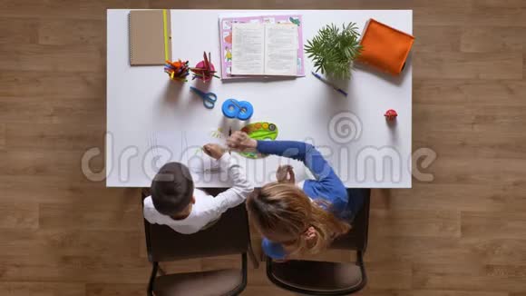 年轻的母亲和儿子一起画画给孩子解释坐在桌子后面男孩在做作业上面的木球拍视频的预览图