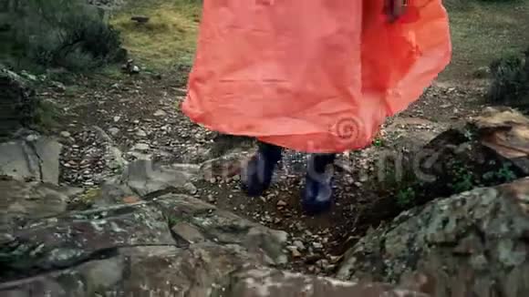穿着橙色雨衣和蓝色雨靴的旅游妇女在石山上升起穿上靴子视频的预览图