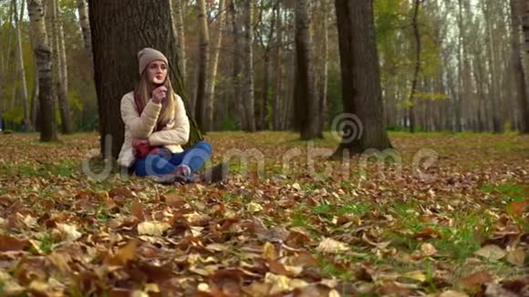 女孩独自坐在秋天的公园里在一棵树下的地上视频的预览图