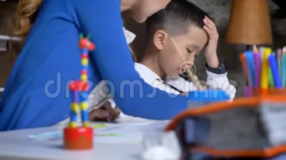 亚洲年轻的母亲给儿子解释帮孩子做作业坐在桌子后面亚洲男孩抱着他的头看书视频的预览图