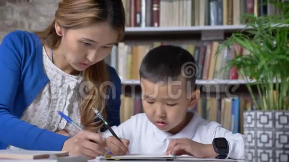 亚洲年轻的母亲和儿子一起写作女人帮助孩子坐在桌子后面亚洲孩子做家庭作业书架视频的预览图