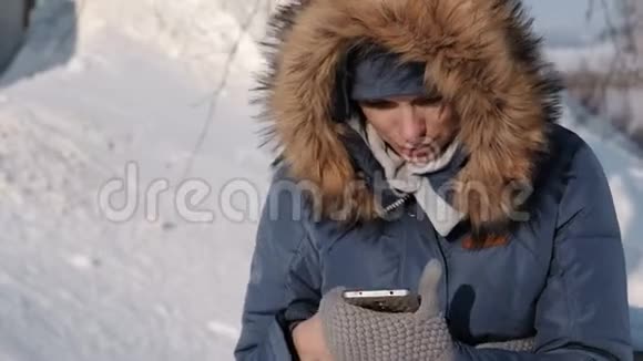 一个穿着蓝色羽绒服的特写女人在冬季公园的手机里写信息前视图视频的预览图