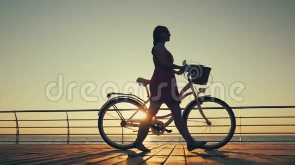 日出或日落时穿着老式自行车在海边的木堤上行走的年轻女子剪影视频的预览图