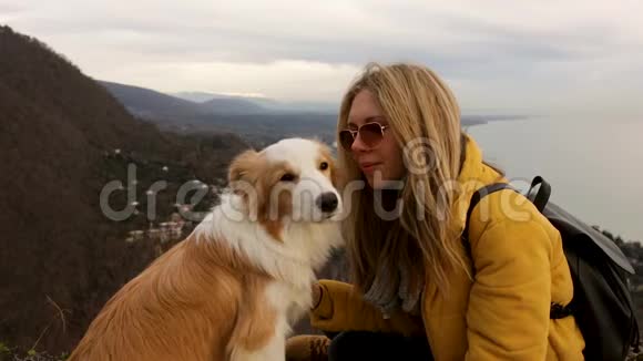 旅行美丽的女孩带着眼镜亲吻和拥抱狗边境牧羊犬在山顶海景城市视频的预览图