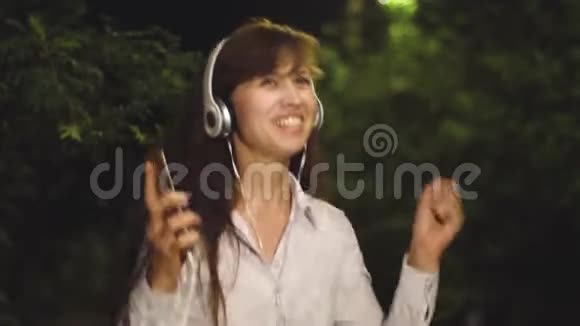 戴着耳机和触摸电话的女孩听着音乐在灯笼的照耀下微笑着在夜城跳舞视频的预览图