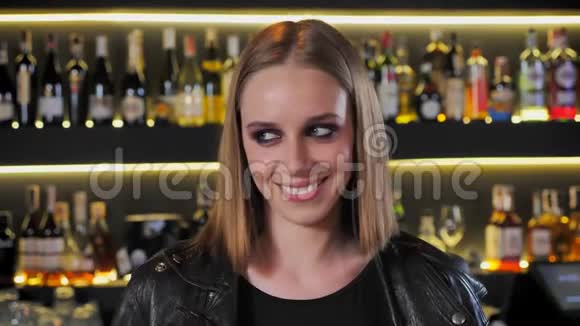 年轻迷人的女人穿着黑色夹克浓妆艳抹看着镜头笑着笑着酒吧背景视频的预览图