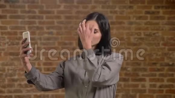 年轻迷人的女人棕色短发用电话和触摸头发自拍严肃砖墙背景视频的预览图