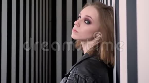 年轻漂亮的女人穿着黑色夹克靠在墙上转身看着相机条纹墙视频的预览图