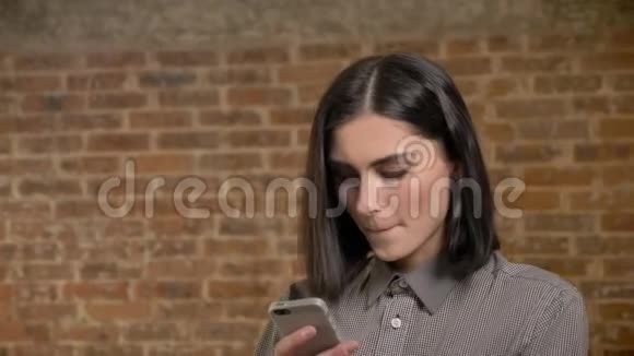 年轻漂亮的女人在电话里发短信咬着嘴唇集中的砖墙背景的肖像视频的预览图