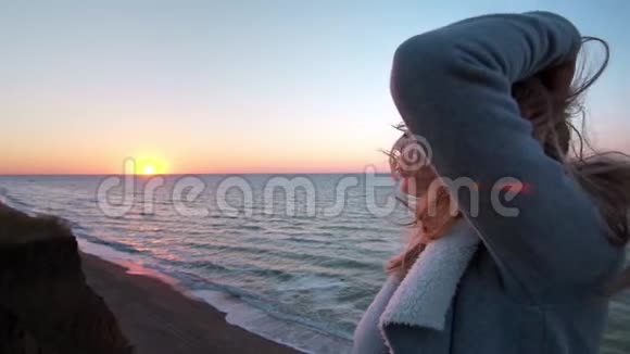 美丽的白种人卷曲的金发孤独的年轻女子在日出或日落的海洋海岸收集她的头发凌乱视频的预览图