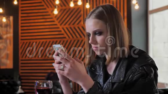年轻漂亮的女人穿着黑色夹克浓妆艳抹地在电话里发短信集中咖啡馆背景视频的预览图