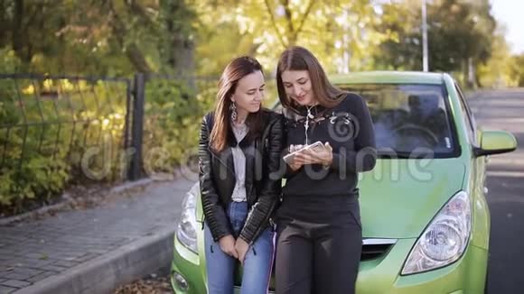 两个女人站在一辆停着的汽车的引擎盖上互相交谈从电话里互相展示信息和照片视频的预览图