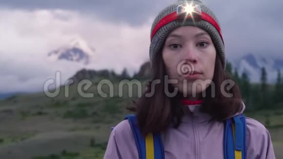 一个年轻的女人带着背包和前照灯在攀爬前看着相机背景中的雾山视频的预览图