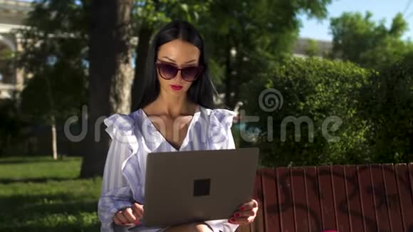 时尚的深色头发戴着太阳镜坐在公园的长凳上在笔记本电脑上打字视频的预览图