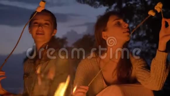两个白种人年轻的女孩晚上坐在大自然的炉火旁吃着沼泽看着明火思考着视频的预览图