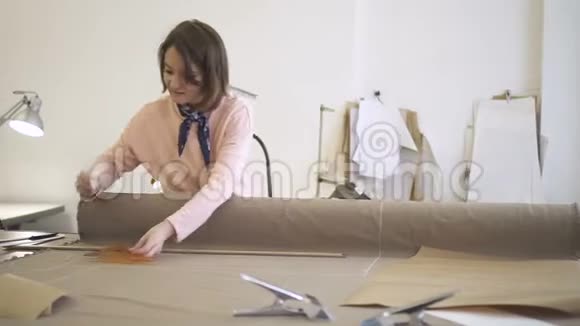 年轻的女时装设计师正在工作用粉笔和尺子在纺织品上做标记站在旁缝纫视频的预览图