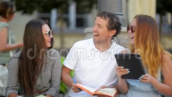 坐在长凳上的三个微笑的学生在一起聊天和学习的户外肖像两个女孩和视频的预览图