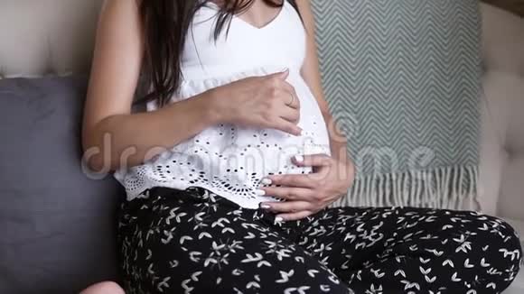 美丽的孕妇坐在沙发上抚摸着她的腹部看着漂亮的相机的肖像视频的预览图