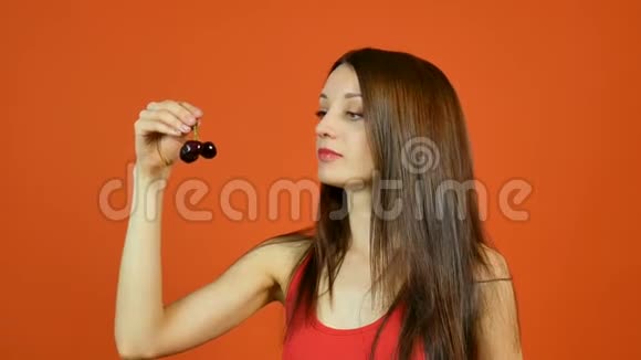 微笑的年轻女性在工作室橙色背景下吃两个暗红色樱桃的肖像健康营养视频的预览图