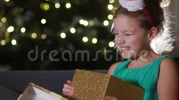 可爱的女孩为圣诞礼物而高兴女生正坐在新年树对面的沙发上抱着一个盒子视频的预览图