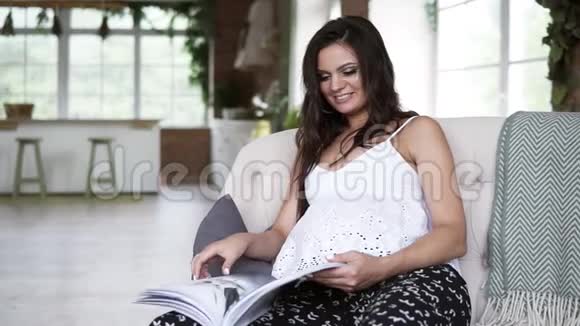 年轻孕妇坐在沙发上布吕内特女士长发阅读杂志放松休闲时间前线视频的预览图