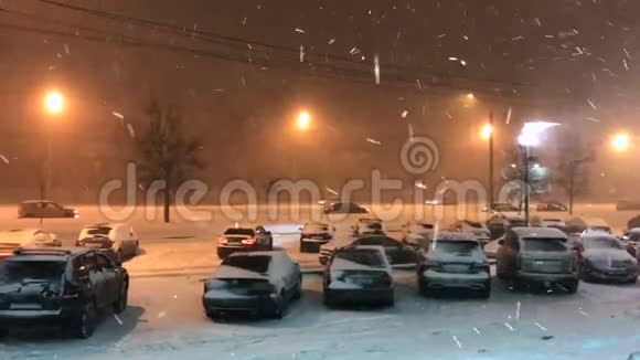 傍晚在大雪下汽车在灯光下缓慢地沿着道路行驶白雪覆盖的汽车停在停车场视频的预览图