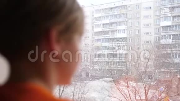 阳光明媚的冬天白天一个年轻人站在窗户旁边看着飘落的雪视频的预览图