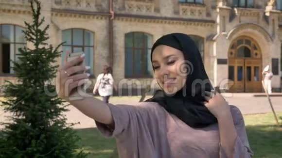 年轻漂亮的穆斯林女孩戴着头巾正在智能手机上自拍在夏天的白天就像一个标志微笑着视频的预览图