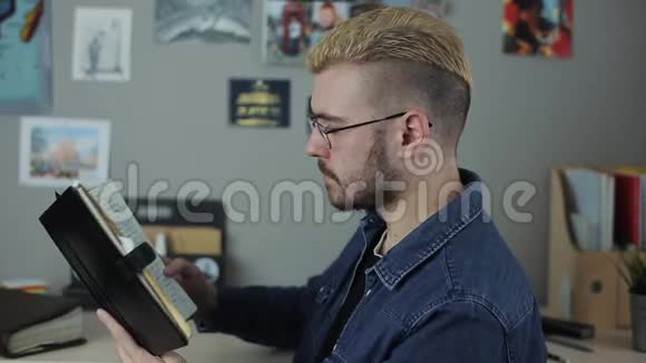 休闲成熟的男学生发型时尚留着黄色头发坐在家里的办公室里读一本棕色的大书视频的预览图