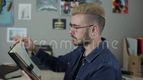 休闲成熟的男学生发型时尚留着黄色头发坐在家里的办公室里读一本棕色的大书视频的预览图
