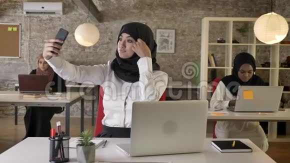 三个戴着头巾的年轻穆斯林妇女在现代办公室工作而其他穆斯林妇女则用智能手机自拍视频的预览图