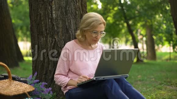 漂亮的女性养老金领取者坐在公园里手提电脑和网上购物视频的预览图