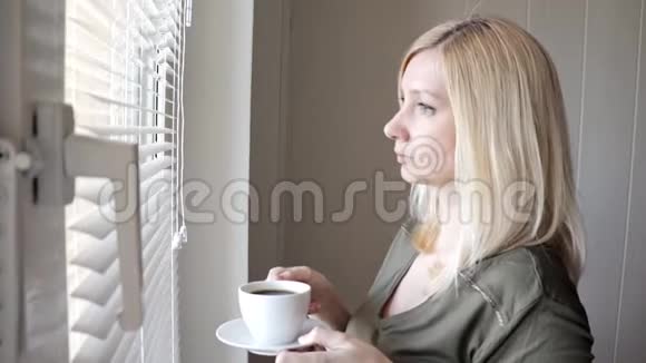 一个年轻的忧郁的美丽的金发女人早上站在窗户附近戴着百叶窗喝着咖啡视频的预览图